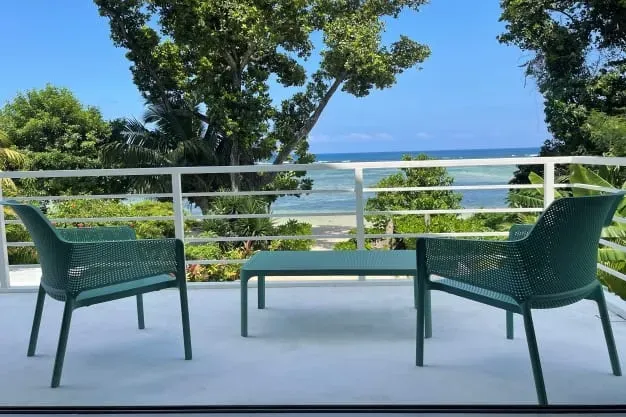 View from a balcony overlooking a tropical beach with lush greenery, blue ocean, and clear sky, featuring outdoor seating with two chairs and a table at Lyla Beach Villa.