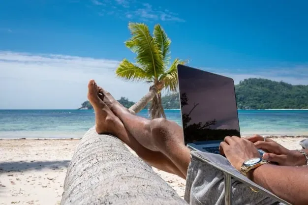 A person relaxing on a tropical beach while working on a laptop, with palm tree and ocean view in the background.