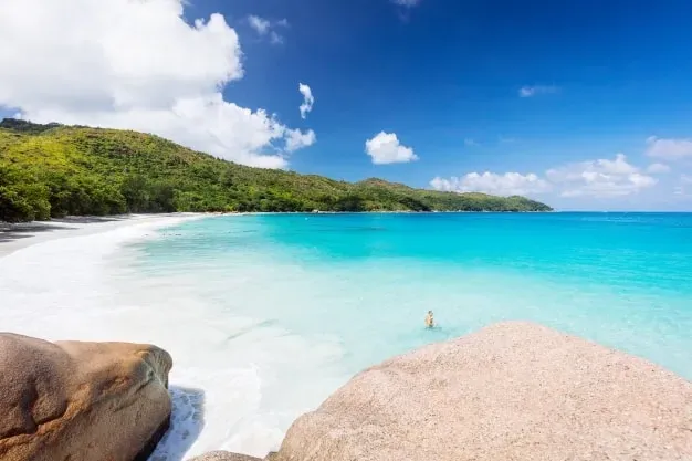 Tropical beach with clear turquoise waters, white sand, and lush green hills in the background.
