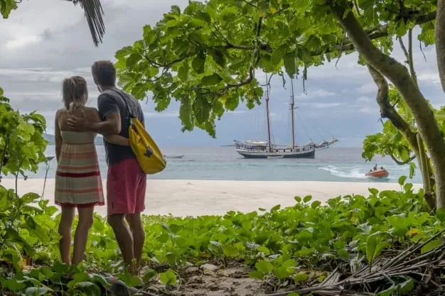 beach lovers on the beach enjoying ocean view, surrounded by lush greenery, with a sailing boat in the background.